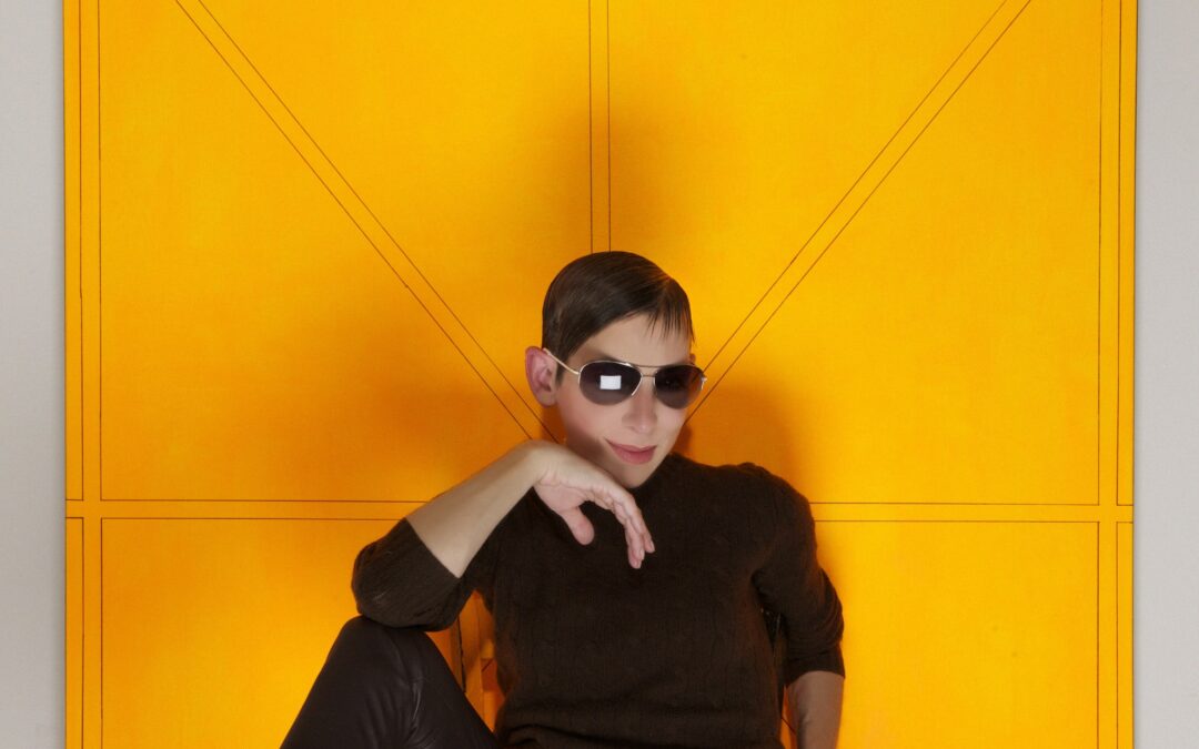 Ivy Brown in black leather outfit with dark sunglasses posed against a bright yellow backdrop