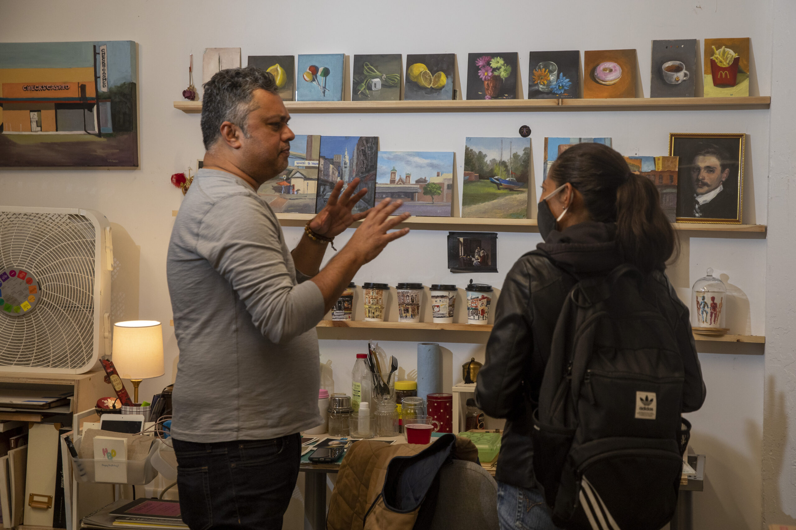 artist Debasish Sinha discusses his artwork with a visitor in his studio with small still life and landscape paintings on the wall in the background