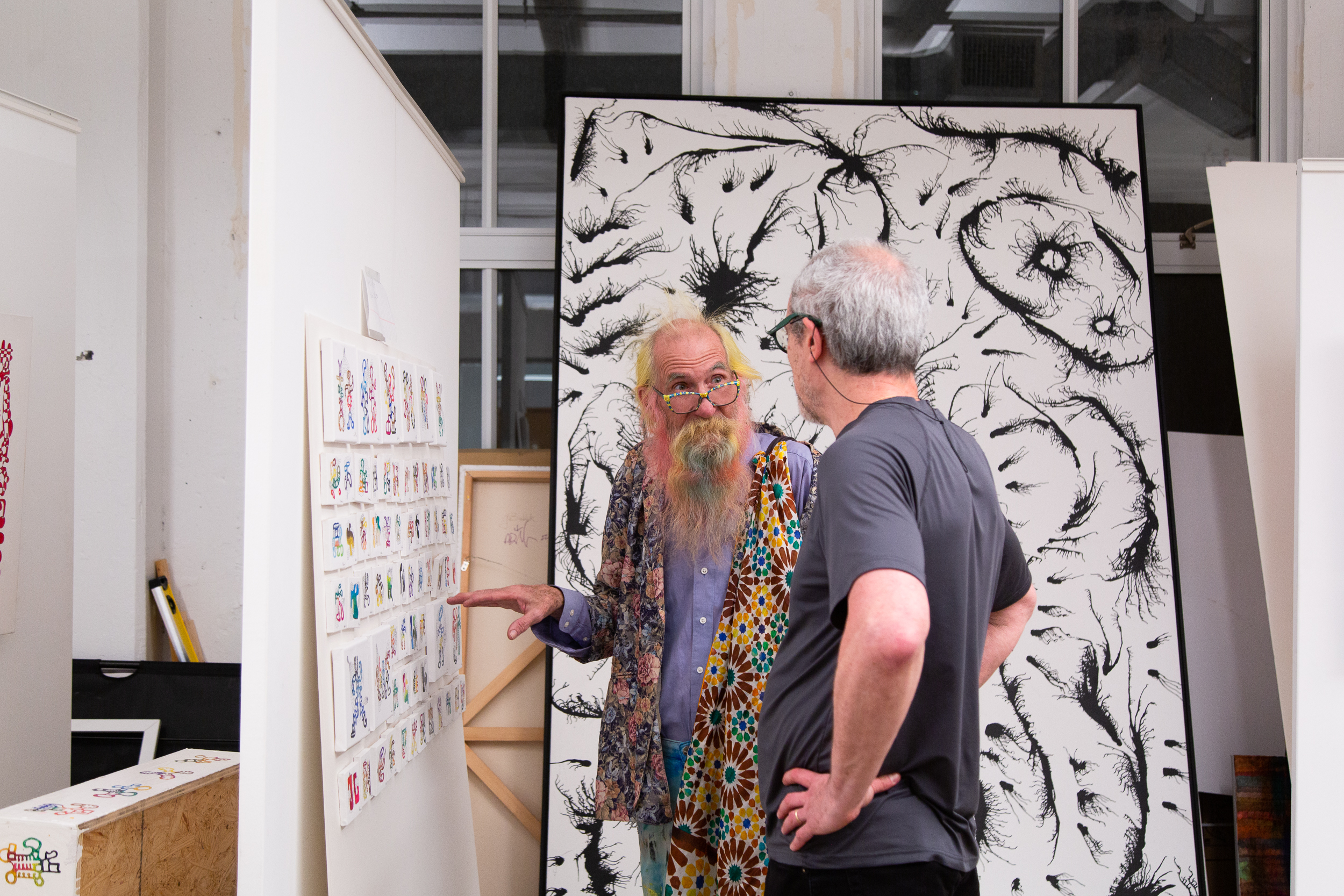 artist Debasish Sinha discusses his artwork with a visitor in his studio with small still life and landscape paintings on the wall in the background