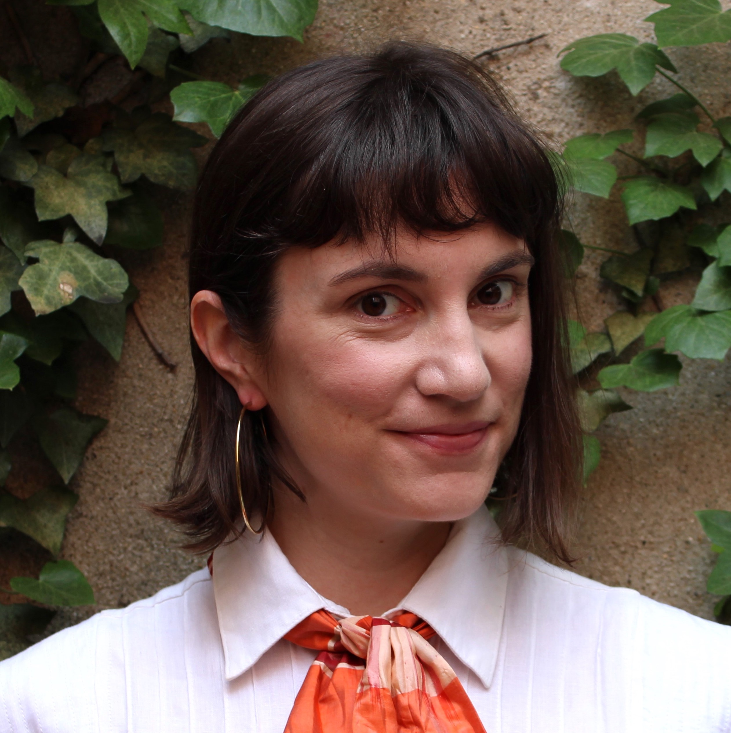 head shot of a woman wearing a white collared shirt with an orange scarf and with brown straight hair just above her shoulders with bangs and her face at an angle